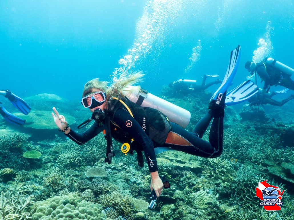 A female scuba diver with braided hair hovering in a horizontal trim position just above a vibrant coral reef.