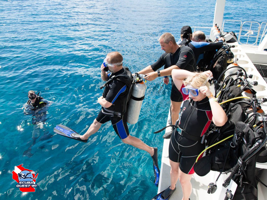 A female scuba diver giving a peace sign while swimming over a coral reef during a guided group dive.